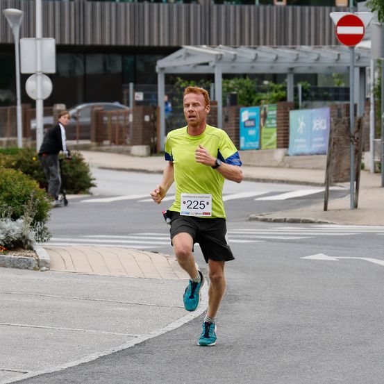 Ein Mann in einem grünen T-Shirt und schwarzen Shorts läuft auf der Straße. Er hält sich am Bauch, vielleicht fühlt er sich unwohl. Eine Person in einer schwarzen Jacke fährt im Hintergrund mit dem Fahrrad. Es gibt auch Pflanzen, ein Gebäude und ein Schild in der Gegend.