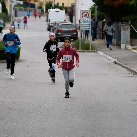 Läufer nehmen auf einer Straße mit Geschwindigkeitsbegrenzungsschild an einem Rennen teil, tragen nummerierte Trikots und Turnschuhe. Zuschauer beobachten vom Bürgersteig aus.