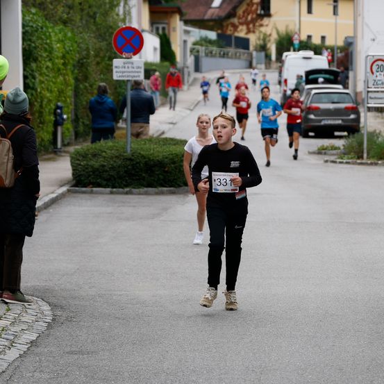 Eine Straßenszene während eines Rennens zeigt einen Jungen mit der Nummer 331, der läuft, gefolgt von anderen. Zuschauer sind an der Seite. Ein Schild zeigt eine Einfahrtsverbotszone an.