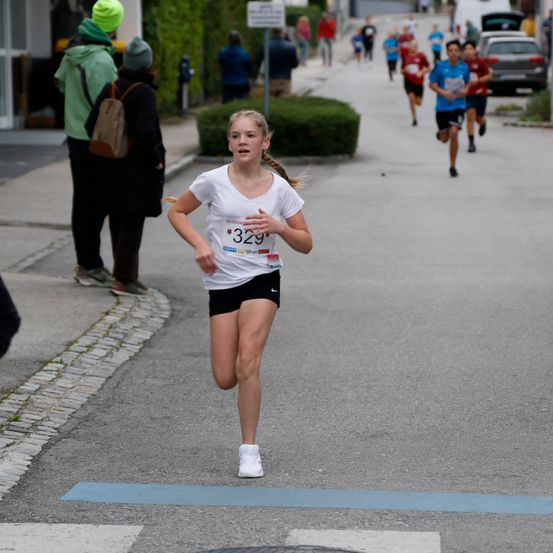 Ein Mädchen mit einem Nummer-329-Shirt läuft bei einem Marathon auf einer Straße, hinter ihr mehrere Läufer und Zuschauer am Straßenrand.