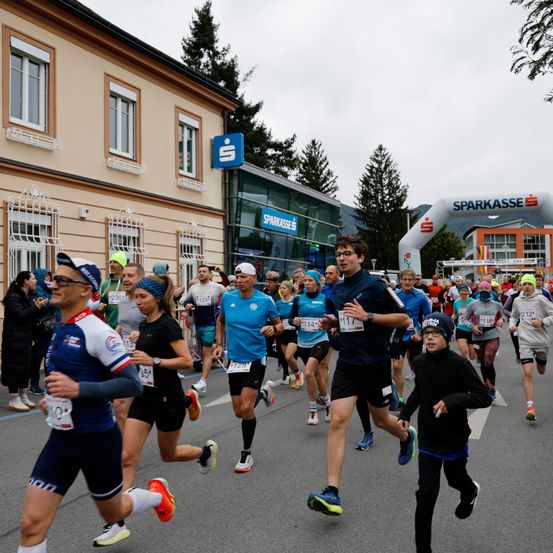 Eine Gruppe von Läufern nimmt an einem Marathon teil. Sie laufen auf der Straße vor einem Gebäude mit einem Sparcasse-Schild. Die Läufer tragen verschiedene Sportbekleidung, und einige haben Nummern auf ihren Trikots.