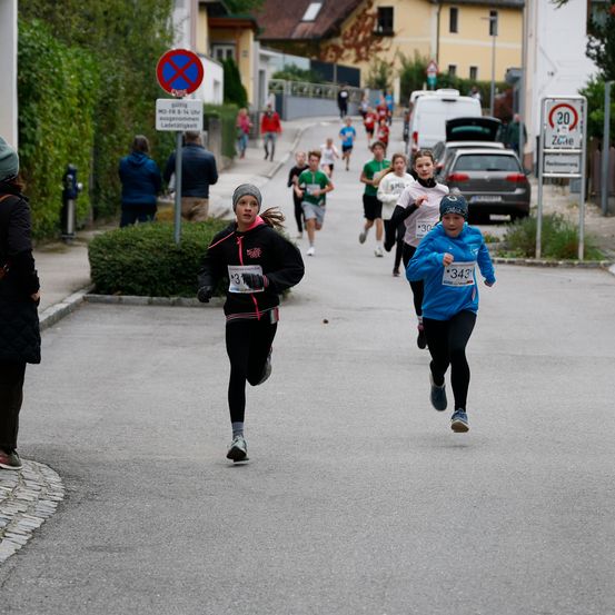 Eine Gruppe von Kindern und Erwachsenen nimmt an einem Laufevent teil. Sie laufen eine Straße hinunter, einige tragen nummerierte Startnummern. Im Hintergrund stehen mehrere Personen in der Nähe eines geparkten Autos und Straßenschildern.