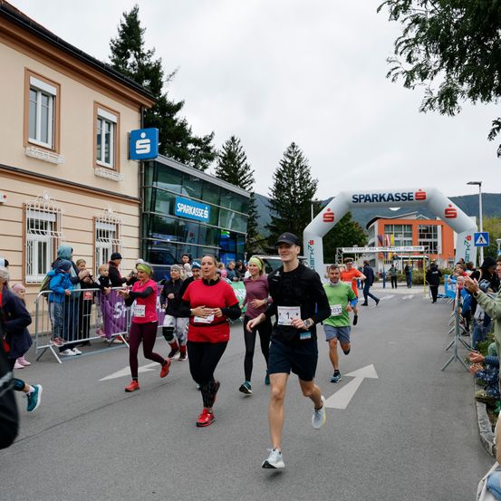 Läufer nehmen an einem Marathon auf einer Straße teil. Zuschauer jubeln an den Seitenlinien. Die Veranstaltung wird von Sparkasse gesponsert.