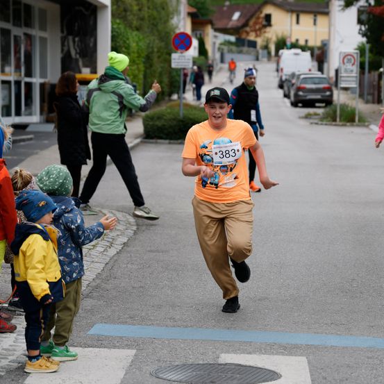 Ein Junge läuft in einem Marathon auf der Straße. Zuschauer jubeln an der Seite. Der Junge trägt die Nummer 383 auf seinem Shirt.