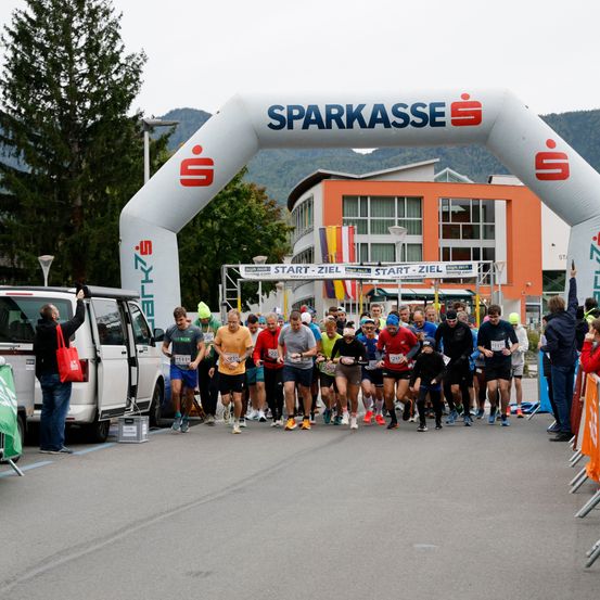 Läufer laufen durch einen weißen Bogen mit der Aufschrift 'Sparkasse' und beginnen ein Rennen. Zuschauer stehen herum und beobachten. Die Straße ist leer.