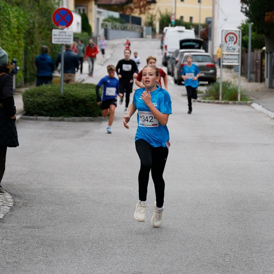 Ein junges Mädchen in einem blauen Shirt und schwarzen Hosen läuft bei einem Rennen. Sie trägt weiße Turnschuhe und hat die Nummer 342 auf ihrem Shirt. Andere Läufer nehmen ebenfalls am Rennen teil.