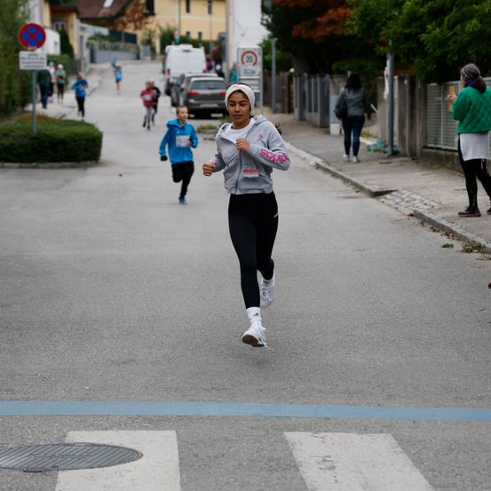 Eine Frau läuft während eines Rennens auf einer Straße, gefolgt von einem Jungen. Andere Teilnehmer, Zuschauer und ein Zaun sind im Hintergrund zu sehen.