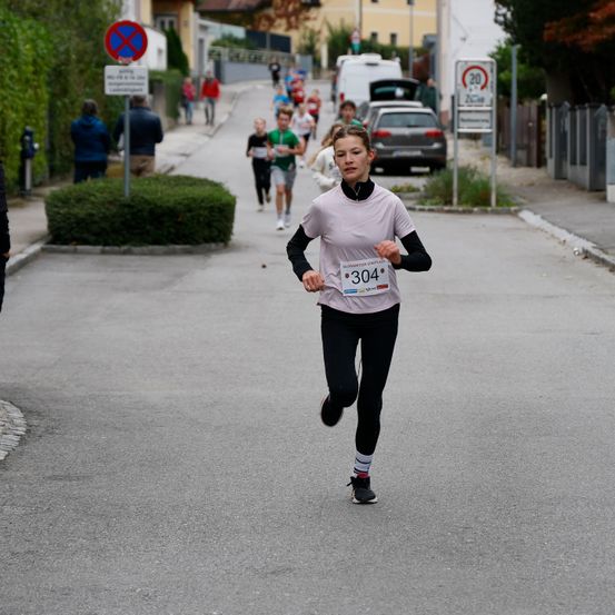 Eine Frau mit der Nummer 304 auf ihrem Shirt läuft einen Marathon. Hinter ihr laufen mehrere Menschen, und der Weg hat Schilder und geparkte Autos am Straßenrand.