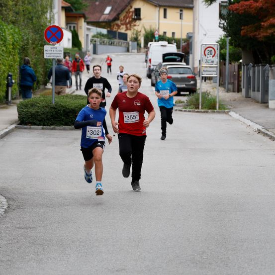 Eine Gruppe von Kindern läuft auf der Straße bei einem Rennen. Ein Kind trägt ein blaues Shirt mit der Nummer 302, während ein anderes Kind in Rot die Nummer 355 trägt.