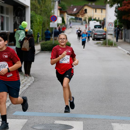 Läufer nehmen an einem Rennen auf einer Straße teil. Ein junger Mensch mit Brille und Nummer 357 läuft vorneweg. Zuschauer und geparkte Autos säumen die Straße.