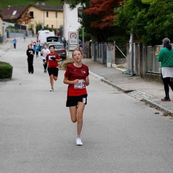 Eine Frau in einem roten Shirt mit der Nummer 361 läuft bei einem Marathon. Hinter ihr folgen weitere Läufer. Die Straße ist von Gebäuden und Bäumen gesäumt. Eine Frau am Bürgersteig beobachtet.