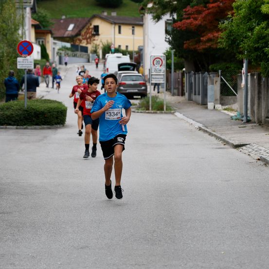Ein Straßenrennen, bei dem ein Läufer mit der Nummer 334 führt, gefolgt von anderen, an einem sonnigen Tag mit Gebäuden und Bäumen im Hintergrund.
