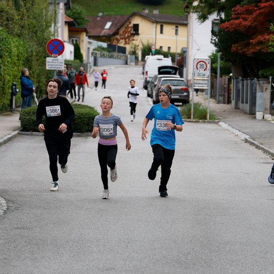 Eine Gruppe junger Menschen läuft in einem Rennen. Drei von ihnen sind in Führung. Sie laufen auf einer Straße. Im Hintergrund sind geparkte Autos, Gebäude und Bäume.