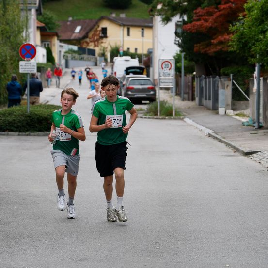 Zwei junge Jungen nehmen an einem Marathon auf einer asphaltierten Straße teil. Sie laufen Seite an Seite mit Nummern auf ihren Trikots. Dahinter folgen einige andere Läufer. Die Straße ist gesäumt von Gebäuden und Bäumen.