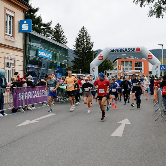 Läufer nehmen an einem Marathon teil, mit Zuschauern an der Seite. Die Route ist mit einem weißen Pfeil markiert. Die Veranstaltung wird von Sparkasse gesponsert.