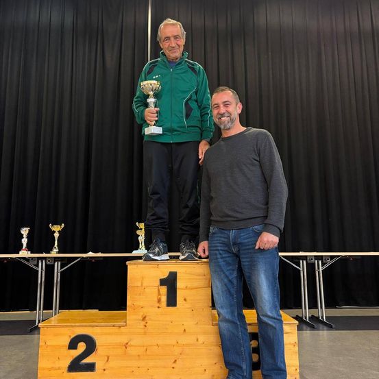 Ein älterer Mann steht mit einer Trophäe in der Hand auf einem Podest, neben einem jüngeren Mann. Dahinter stehen mehrere Trophäen auf einem Tisch und ein schwarzer Vorhang.