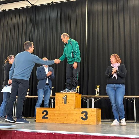 Ein Podium mit drei Stufen, der Gewinner des ersten Platzes auf der obersten Stufe, der einen Händedruck von einem Mann erhält, während zwei Frauen und ein Mann applaudieren.