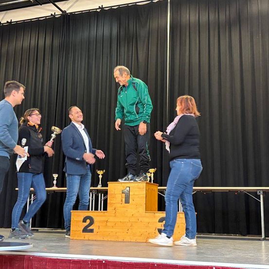 Ein Mann steht auf einem Podium mit dem Fuß auf der ersten Stufe, während andere Trophäen halten. Sie sind in einem Raum mit schwarzen Vorhängen.