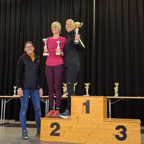 Drei Frauen stehen auf einem Podest und halten Trophäen. Sie tragen lässige Sportkleidung. Das Podest hat die Nummern 1, 2 und 3. Hinter ihnen sind auf einem Tisch Trophäen ausgestellt. Ein schwarzer Vorhang dient als Hintergrund.