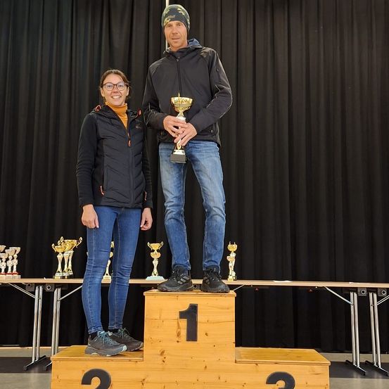 Zwei Personen stehen auf einem Podest mit der Nummer eins und halten eine Trophäe. Sie lächeln und scheinen für ein Foto zu posieren. Dahinter sind auf einem Tisch Trophäen angeordnet. Ein schwarzer Vorhang ist im Hintergrund.