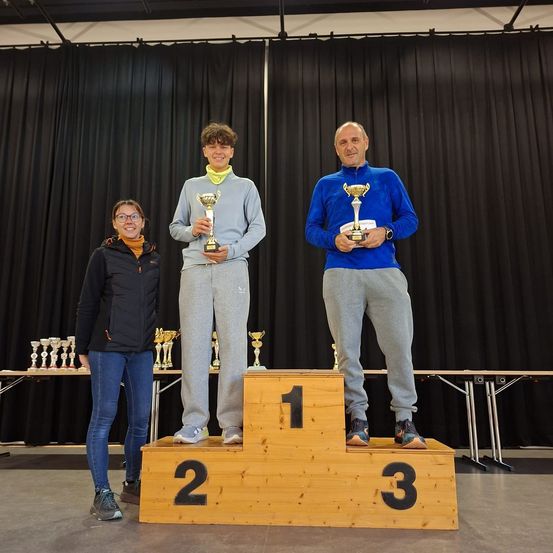 Drei Personen stehen auf einem Podest mit den Nummern 1, 2 und 3 und halten Pokale. Sie sind in einem Raum mit schwarzen Vorhängen und einem Tisch mit Pokalen.