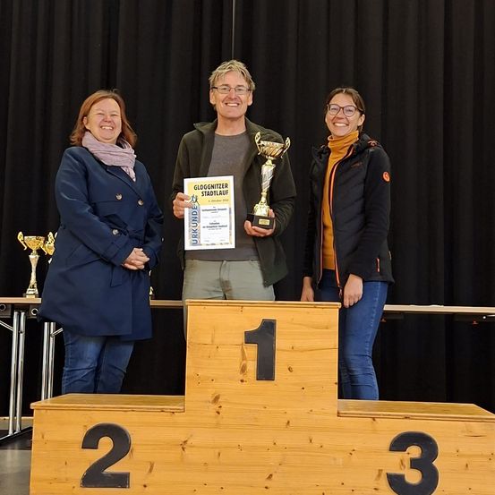 Drei Personen stehen auf einem Podest mit den Nummern eins, zwei und drei. Der Mann in der Mitte hält einen Pokal und ein Zertifikat. Hinter ihnen befindet sich ein schwarzer Vorhang und ein Tisch mit Pokalen.