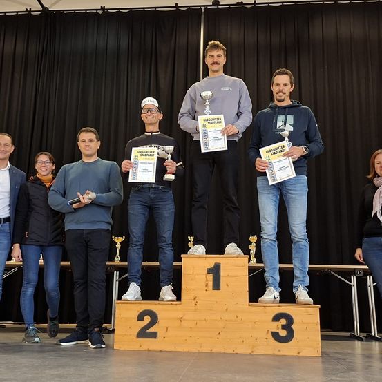 Fünf Männer und eine Frau stehen auf einem Podium mit Trophäen. Sie tragen Winterkleidung. Das Podium ist aus Holz mit den Nummern 1, 2 und 3.