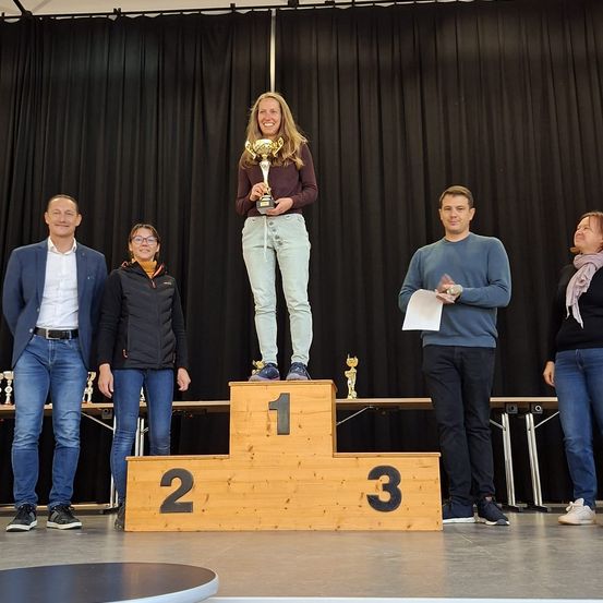 Eine Frau steht auf dem Podest des ersten Platzes und hält einen Pokal in der Hand, während vier Personen um sie herum stehen, eine mit einem Papier. Hinter ihnen steht ein Tisch mit Pokalen. Der Hintergrund besteht aus schwarzen Vorhängen.