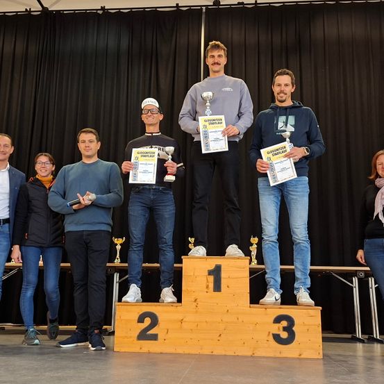 Drei Männer stehen mit Trophäen auf einem Podium, tragen Jacken und Turnschuhe. Sie sind umgeben von anderen, einige halten Trophäen, auf einer Bühne mit schwarzem Hintergrund.