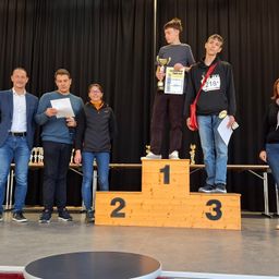 Drei Personen stehen auf einem Podium mit den Nummern 1, 2 und 3 und halten Pokale. Zwei Männer stehen neben ihnen, einer liest ein Papier. Der Hintergrund ist ein schwarzer Vorhang mit Tischen und Stühlen dahinter.
