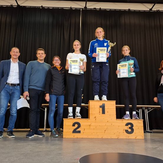 Mehrere Personen stehen auf einem Podium mit Trophäen. Zwei Mädchen stehen in der Mitte, eine Frau rechts und drei Männer links. Dahinter befindet sich ein schwarzer Vorhang.