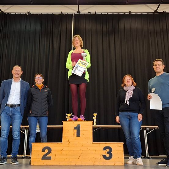 Fünf Personen stehen auf einem Podium mit den Nummern 1, 2 und 3. Die Frau in der Mitte hält eine Trophäe und ein Zertifikat. Hinter ihnen ist ein schwarzer Vorhang teilweise geöffnet.