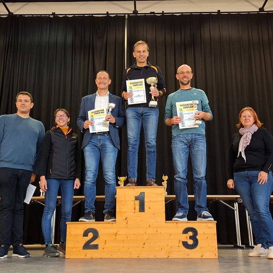 Fünf Personen stehen auf einem Podest mit drei Ebenen. Die oberste Ebene hat die Nummer 1, die mittlere 2 und die unterste 3. Jede Person hält einen Pokal. Der Hintergrund besteht aus schwarzen Vorhängen.