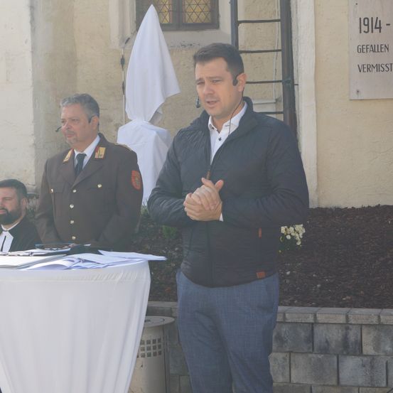Ein Mann in einer schwarzen Jacke steht vor einem Podium, möglicherweise hält er eine Rede. Hinter ihm steht ein Mann in Uniform in der Nähe eines weißen Tuchs und eines Schildes. Ein dritter Mann sitzt an einem Tisch mit Papieren.