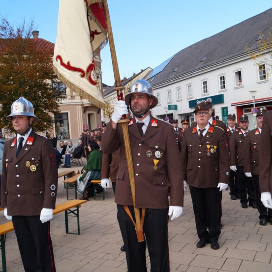 Eine Gruppe uniformierter Männer, die eine Fahne halten, marschiert in einer Parade, mit mehreren Personen, die auf Bänken hinter ihnen sitzen.