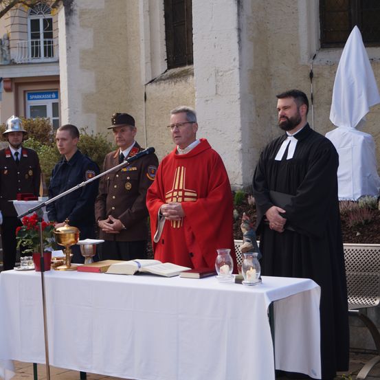 Eine religiöse Zeremonie findet vor einer Kirche statt. Ein Priester steht an einem Tisch, flankiert von zwei anderen, während uniformierte Männer hinter ihnen stehen.