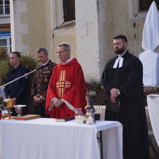 Ein Priester in einer roten Robe steht an einem Tisch mit Kerzen und Büchern, während andere zusehen. Sie befinden sich in der Nähe eines Gebäudes mit einem weißen Sonnensegel.