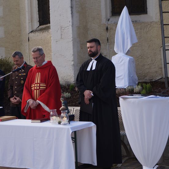 Zwei Männer in religiösen Gewändern stehen vor einem Tisch. Einer in einem roten Gewand und der andere in einem schwarzen Gewand. Ein weißer Stoff bedeckt einen nahen Tisch.