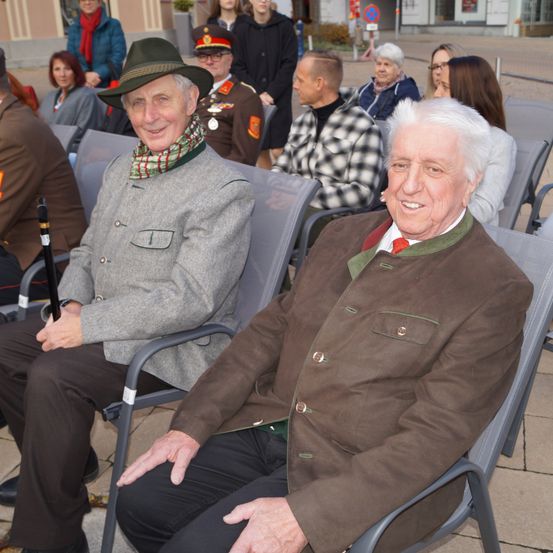 Zwei ältere Männer sitzen nebeneinander in grauen Stühlen, beide in warmen Jacken. Der Mann links trägt eine graue Jacke und einen Schal. Hinter ihnen sitzen Menschen in Winterkleidung in Reihen.