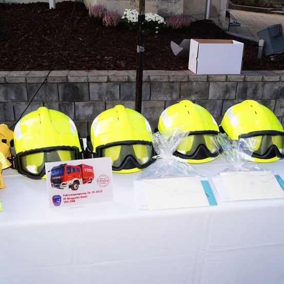 Vier gelbe Helme und Schutzbrillen sind auf einem Tisch ausgestellt, neben einem weißen Tischtuch und einer Feuerwehr-Karte. Hinter dem Tisch befindet sich eine Steinmauer mit Pflanzen und einer weißen Box.
