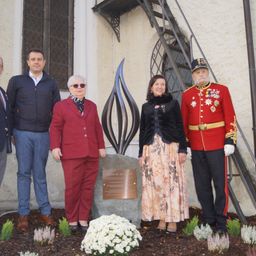Fünf Personen stehen neben einem Denkmal, gekleidet in formelle Kleidung. Ein Mann in einem roten Militäruniform steht rechts, während eine Frau in Rot links steht. Ein Denkmal steht dahinter.