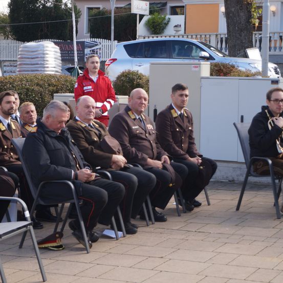 Eine Gruppe uniformierter Männer sitzt auf Stühlen, möglicherweise für eine Zeremonie. Ein Mann in einem roten Jackett steht hinter ihnen. Im Hintergrund ist ein Auto geparkt.