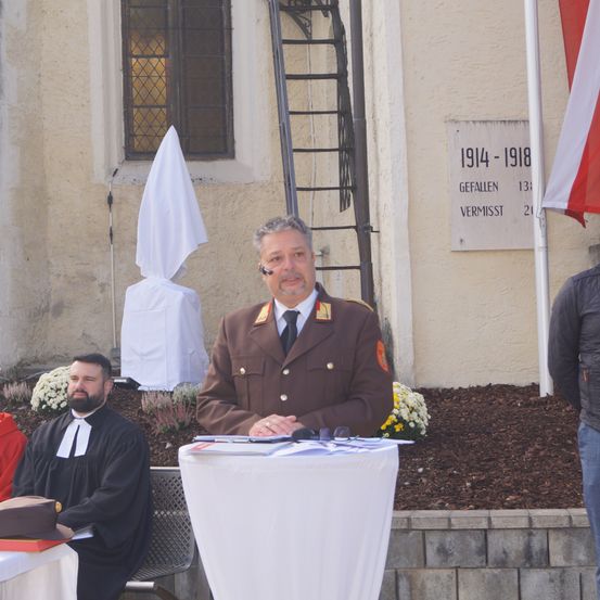 Ein Mann in Uniform spricht an einem Podium vor einem Denkmal. Ein Priester sitzt hinter ihm. Dahinter steht ein Gebäude mit einer Flagge und einer Gedenktafel.