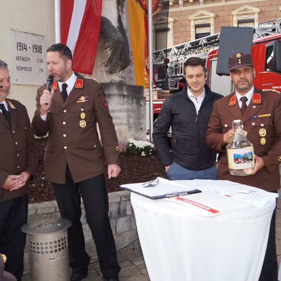 Drei Männer in Uniformen stehen um einen Tisch mit Papieren. Einer hält eine Flasche und spricht in ein Mikrofon. Dahinter steht ein Feuerwehrwagen und ein Denkmal mit den Jahren 1914-1918.