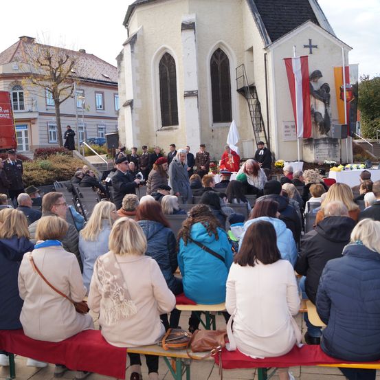 Eine Menschenmenge versammelt sich vor einer Kirche, einige auf Bänken sitzend, andere stehend. Ein Podium ist in der Nähe des Kircheneingangs aufgestellt, und Flaggen sind an der Seite der Kirche angebracht.