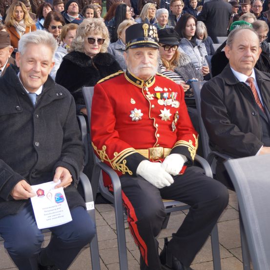 Ein Mann in einem roten Uniform mit Medaillen sitzt zwischen drei anderen. Einer hält ein Papier mit Text. Die Menge ist warm gekleidet.