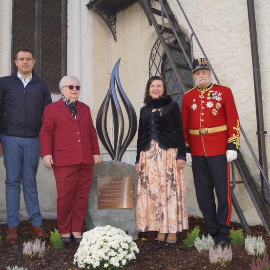 Vier Personen stehen neben einer flammenförmigen Skulptur in einem Garten und lächeln für ein Foto. Dahinter befindet sich eine Steinplatte mit Text.