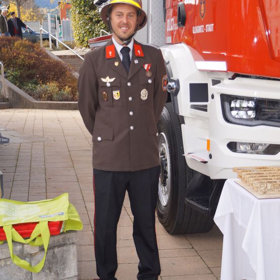 Ein Feuerwehrmann in Uniform steht vor einem Feuerwehrwagen. Hinter ihm befindet sich eine grüne Tasche und ein Tisch mit einem weißen Tuch und verschiedenen Gegenständen.