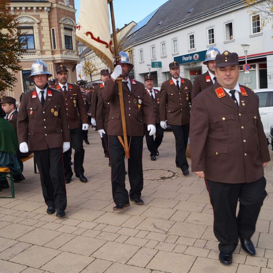 Eine Gruppe uniformierter Männer marschiert mit einer Fahne in einem Umzug und trägt Helme und Handschuhe. Sie befinden sich auf einem Ziegelweg vor Gebäuden.