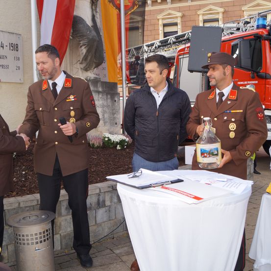 Drei Männer stehen vor einem Gedenkstein mit Fahnen und einem Feuerwehrauto im Hintergrund. Ein Mann hält ein Mikrofon, ein anderer eine Flasche und der dritte gibt einem Mann in einem braunen Uniform die Hand.